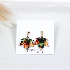 May include: A pair of gold dangle earrings with a watercolor-style maple leaf design in shades of green, orange, red, and yellow. The earrings are on a white card with the text "grace + joy collective".