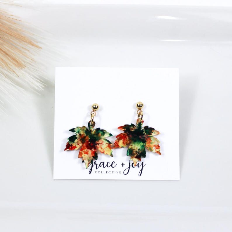 May include: A pair of gold dangle earrings with a watercolor-style maple leaf design in shades of green, orange, red, and yellow. The earrings are on a white card with the text "grace + joy collective".