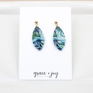 May include: A pair of leaf-shaped earrings with a blue, green, and white watercolor design. The earrings have gold-colored posts and are displayed on a white card with the text "grace + joy collective" in black script.