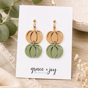 Peut inclure: Une paire de boucles d'oreilles pendantes composées de deux formes de citrouilles superposées, l'une beige et l'autre vert sauge, suspendues à des clous dorés. Elles sont présentées sur une carte blanche avec le texte grace + joy collective.