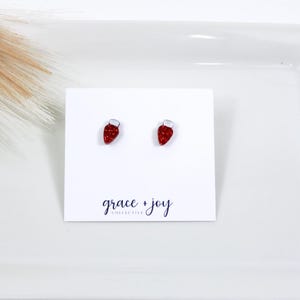 May include: A pair of red glitter Christmas light bulb stud earrings with white tops. The earrings are displayed on a white card with the text "grace + joy collective".