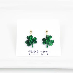 May include: A pair of green shamrock earrings with gold-colored posts. The earrings are on a white card with the text "grace + joy" in a script font. The shamrocks have a glittery effect.