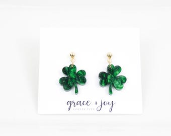 Könnte beinhalten: Ein Paar grüne Kleeblatt-Ohrringe mit goldfarbenen Pfosten. Die Ohrringe befinden sich auf einer weißen Karte mit dem Text "grace + joy". Die Kleeblätter haben einen Glitzereffekt. Ideal für St. Patrick's Day.