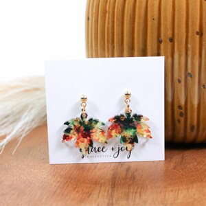 May include: A pair of gold dangle earrings with a colorful fall leaf design. The leaves are a mix of red, orange, yellow, and green. The earrings are on a white card with the text "grace + joy collective".