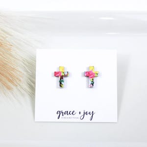 May include: A pair of floral cross stud earrings. The cross is yellow with a pink and white floral pattern. The earrings are on a white card with the text "grace + joy collective".