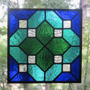 May include: A square stained glass panel with a geometric design. The panel features a symmetrical pattern of blue, teal, and green glass pieces, outlined in black. Small, square, textured clear glass accents are interspersed throughout the design. The panel is suspended by a chain.