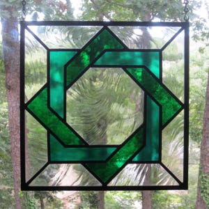 May include: Square stained glass panel with a geometric design. The panel features a central green knot design surrounded by clear and green glass sections, all framed by black lines. The panel is hanging in front of a blurred outdoor background.