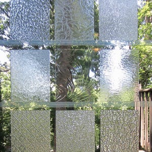 May include: A grid of nine rectangular glass panels arranged in three rows and three columns. Each panel features a distinct textured or patterned surface, including floral, wavy, pebbled, and geometric designs, all made of clear glass.