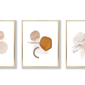 Op de afbeelding: Set van drie abstracte kunstprints in gouden lijsten. Elke print heeft een andere opstelling van bruine, beige en witte vormen op een witte achtergrond.