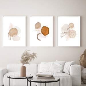 Op de afbeelding: Drie abstracte aquarelprints in witte frames met bruine en beige vormen en patronen. De prints hangen aan een witte muur boven een witte bank en twee zwarte salontafels.