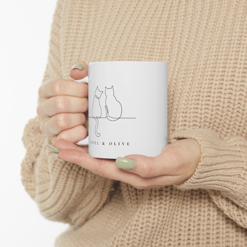Pu&ograve; includere: Tazza in ceramica bianca con un disegno a linea nera di due gatti seduti uno accanto all'altro. Il testo "Axel & Olive" &egrave; stampato sul fondo della tazza.