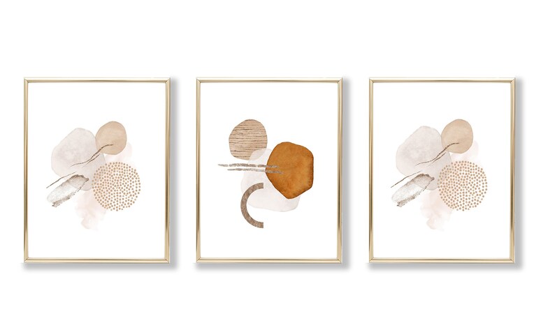 Op de afbeelding: Set van drie abstracte kunstprints in gouden lijsten. Elke print heeft een andere opstelling van bruine, beige en witte vormen op een witte achtergrond.