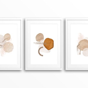 Op de afbeelding: Drie ingelijste prints met abstracte vormen in tinten bruin en beige. De vormen zijn organisch en lijken op cirkels, ovalen en lijnen.