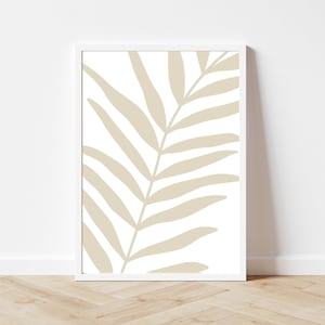 Op de afbeelding: Een ingelijste print van een beige palmblad op een witte achtergrond. De print is in een wit frame en ligt op een lichte houten vloer.