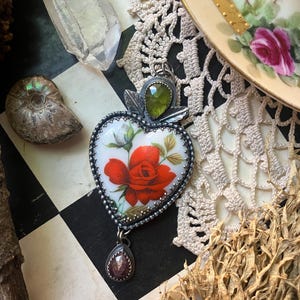 Puede incluir: Un colgante en forma de corazón con un centro de cerámica blanca que presenta una ilustración de una rosa roja y hojas verdes. El colgante está enmarcado en metal plateado con una piedra verde facetada arriba y una pequeña piedra oscura.