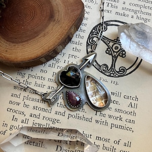 Könnte beinhalten: Eine silberne Halskette mit einem Anhänger in Form eines Pfeils, einem schwarzen Edelstein, einem rosafarbenen Edelstein und einem Stück irisierendem Muschelschalen. Die Halskette wird auf einem Buch mit dem Text "FIG. 113." und "Design" gezeigt. Die Halskette wird auch mit einem klaren Quarzkristall gezeigt.