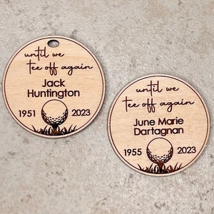 May include: Two round wooden golf memorial tags. Each tag has the phrase "until we tee off again", a name, birth and passing years, and a golf ball graphic. One tag reads "Jack Huntington 1951-2023". The other reads "June Marie Dartagnan 1955-2023".