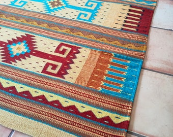 May include: A colorful woven rug with a geometric pattern in shades of blue, red, yellow, and brown. The rug has a fringe on the bottom edge.