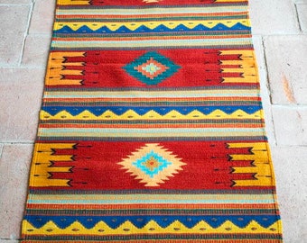 May include: A rectangular, woven rug with a Southwestern design. The rug features a red background with geometric patterns in yellow, blue, and turquoise. The rug has a fringe on both ends and is suitable for a hallway or entryway.