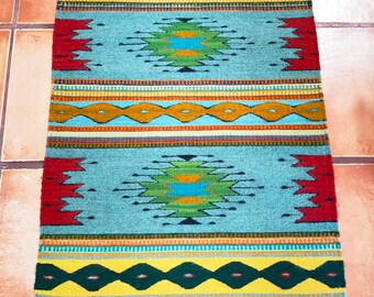 May include: A traditional Native American style rug with a geometric pattern in shades of blue, green, yellow, red, and black. The rug has a fringed edge.