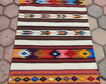May include: A rectangular woven rug with a cream background and colourful geometric patterns. It features horizontal stripes in red, orange, yellow, blue, and brown, with arrow and diamond motifs. The edges have a fringe.