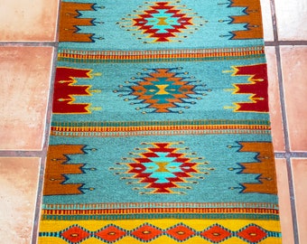 May include: A teal blue rug with a geometric pattern in shades of red, orange, yellow, and blue. The rug has fringe on the ends.