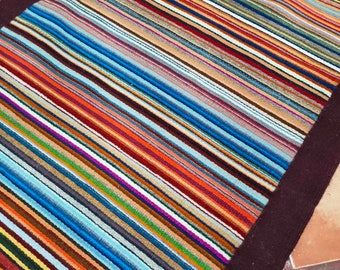 May include: A colorful striped rug with a dark brown border. The rug is made of woven fabric and has a variety of colors, including red, blue, green, yellow, and orange.