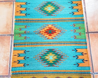 May include: A teal blue rug with a geometric pattern in shades of orange, red, green, and yellow. The rug has a fringe border.