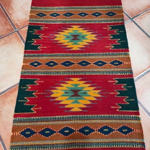 Puede incluir: Una alfombra tejida roja, verde, amarilla y azul con un patrón geométrico. La alfombra tiene flecos en los extremos.