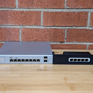 Könnte beinhalten: Ein silberner Netzwerkswitch mit 10 Ports und einem schwarzen Rack-Montagekit. Der Switch hat ein weißes Etikett mit der Aufschrift "Ubiquiti" und das Rack-Montagekit hat ein schwarzes Etikett mit der Aufschrift "Ubiquiti".