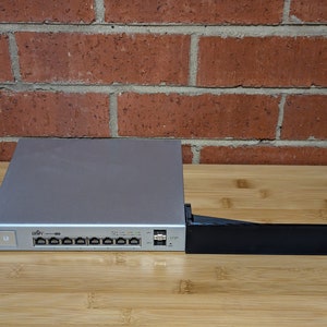 Könnte beinhalten: Ein silberner UniFi-Switch mit acht Ports und einem schwarzen Metall-Rack-Montagekit. Der Switch trägt ein Etikett mit der Aufschrift "UniFi Switch 8 150W".
