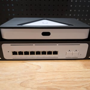 以下が含まれることがあります： 2つの電子機器を収容する黒いラックマウントエンクロージャ。上部のデバイスは白で、黒いロゴと小さな開口部があります。下部のデバイスはシルバーで、複数のポートがあります。ラックには側面に取付穴があります。