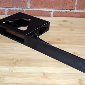 Puede incluir: Soporte negro impreso en 3D con un brazo largo que se extiende hacia la derecha. El soporte tiene una abertura rectangular y una forma triangular en la parte superior. El brazo tiene una superficie lisa y tres agujeros en el extremo.