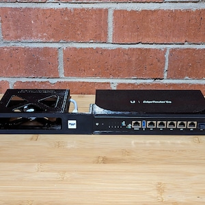 May include: A black network router with the text "Ubiquiti EdgeRouter 6s" on the front. The router has six ethernet ports and a power button. The router is mounted on a black metal rack.