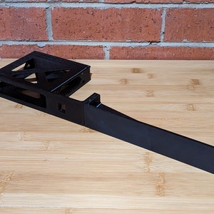 Peut inclure: Une pièce en plastique noire imprimée en 3D avec un long bras droit s'étendant d'une base rectangulaire. La base a une ouverture rectangulaire et un design en treillis.