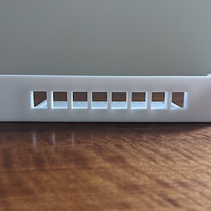 Peut inclure: Support mural en plastique blanc avec dix ouvertures rectangulaires pour la gestion des câbles.