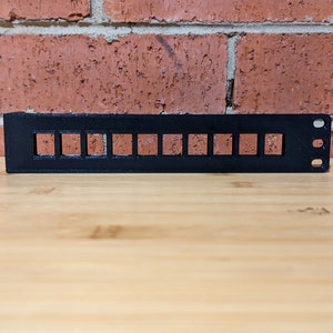 Może przedstawiać: Czarny plastikowy stojak z 12 prostokątnymi otworami. Stojak jest przeznaczony do przechowywania małych przedmiotów, takich jak kable lub przewody.