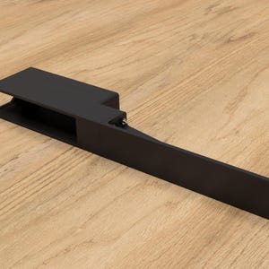 May include: A black metal bracket with a rectangular body and an extended arm. The bracket has a rectangular opening and several mounting holes. The object is set against a light wood grain surface.