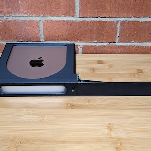 May include: A black plastic stand with a slot for a device, featuring an Apple logo on the front. The stand has a long, thin extension that can be used to mount the device on a wall.