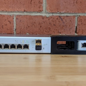 Könnte beinhalten: Ein silberner Netzwerkswitch mit dem Text "UniFi" auf der Vorderseite. Der Switch verfügt über acht Ethernet-Ports und einen einzigen SFP-Port. Der Switch ist auf einem schwarzen Metallrack montiert.