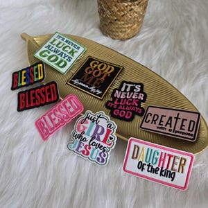 Puede incluir: Un conjunto de parches bordados con frases religiosas e inspiradoras. Los parches presentan textos como "Blessed", "God Got Me" y "Daughter of the King". Están dispuestos sobre una bandeja dorada en forma de hoja.