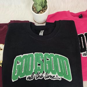May include: Black sweatshirt with the text "GOOSEGOOD all the time" in green and white. A maroon and pink sweatshirt are also visible. The sweatshirts are folded on a white, textured surface.