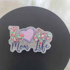 May include: A silver glitter background with the words "Love Mom Life" in blue and pink lettering. The word "Love" is decorated with a floral pattern and a pink heart.