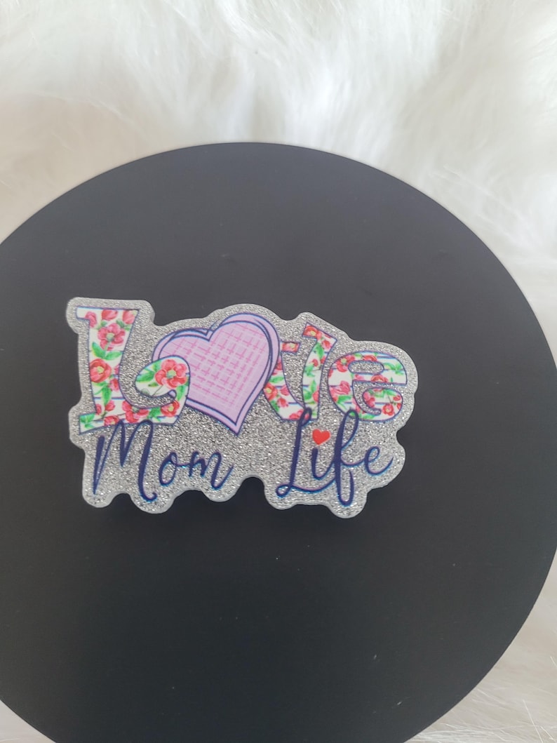 May include: A silver glitter background with the words "Love Mom Life" in blue and pink lettering. The word "Love" is decorated with a floral pattern and a pink heart.