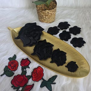 以下が含まれることがあります： 黒と赤のバラのパッチの品揃え。黒いバラは、金の葉の形をしたトレイに配置されています。赤いバラには緑の葉と茎があります。小さな鉢植えの植物が背景にあります。