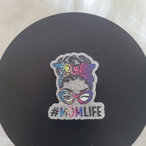 May include: A glittery silver and rainbow coloured sticker with a cartoon woman wearing glasses and a headscarf. The sticker says "#MOMLIFE" in silver glitter.