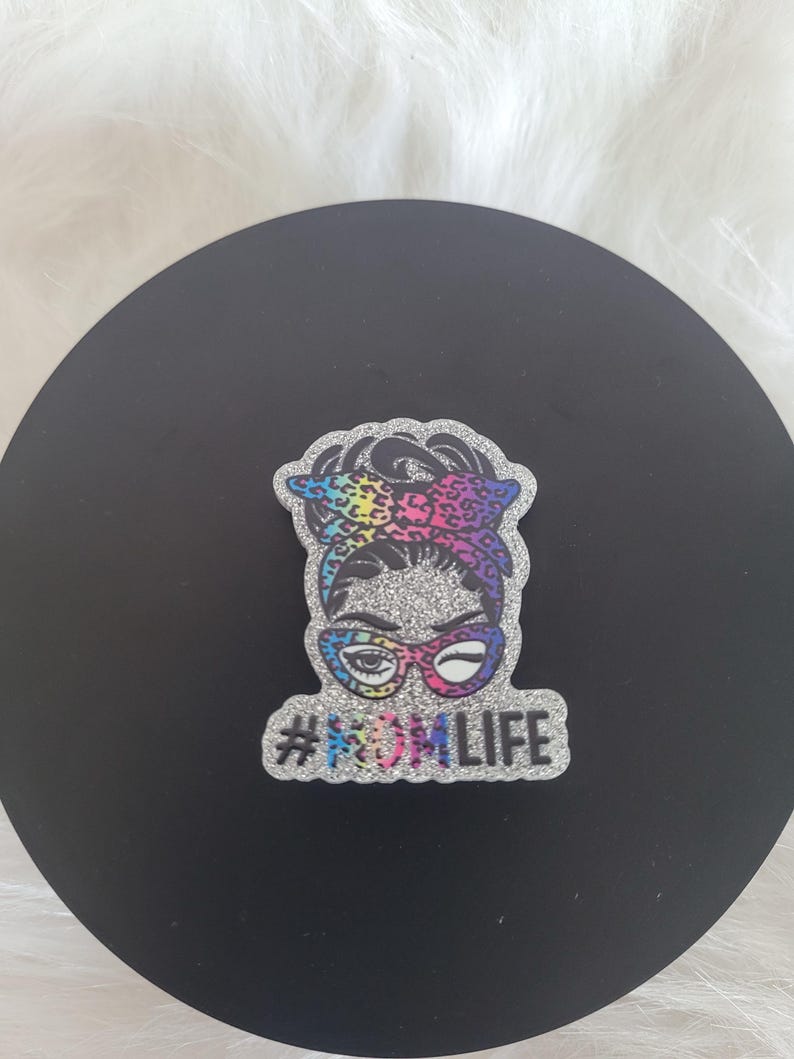 May include: A glittery silver and rainbow coloured sticker with a cartoon woman wearing glasses and a headscarf. The sticker says "#MOMLIFE" in silver glitter.