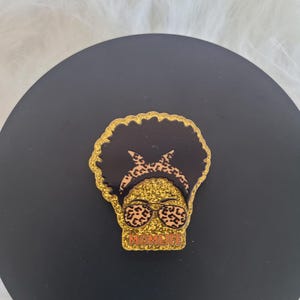 May include: Gold glitter pin featuring a black silhouette of a woman with an afro, sporting leopard print sunglasses and a headband. The text "MOMLIFE" is written in orange beneath the woman's face.