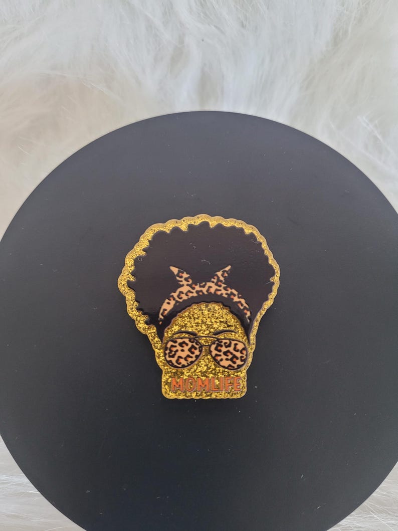 May include: Gold glitter pin featuring a black silhouette of a woman with an afro, sporting leopard print sunglasses and a headband. The text "MOMLIFE" is written in orange beneath the woman's face.