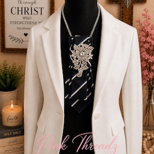 May include: A white blazer worn over a black mannequin, featuring a navy blue and white striped necktie accessory adorned with a large, ornate silver-toned floral brooch encrusted with clear crystals, suspended from a delicate silver-toned chain.